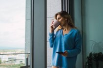 Donna bionda pensierosa che tiene smartphone e tazza di caffè alla finestra — Foto stock
