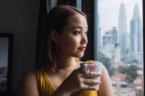 Pensativo asiático mulher com copo olhando através de janela — Fotografia de Stock