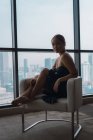 Красива жінка в чорній сукні, що розслабляється в кріслі в квартирі з видом на місто — стокове фото
