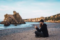 Веселий бородатий чоловік сидить на пляжі і використовує смартфон — стокове фото