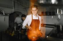 Cocinero haciendo un flambe en la cocina del restaurante con colegas de fondo - foto de stock