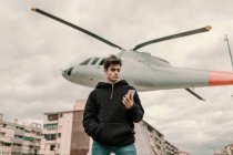 Красивий молодий чоловік стоїть біля пам'ятника вертольоту в місті і використовує смартфон — стокове фото