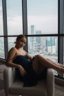 Hübsche Frau in schwarzem Kleid entspannt im Sessel in der Wohnung mit Blick auf die Stadt — Stockfoto