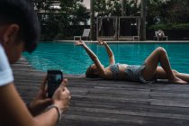 Фотограф фотографирует азиатку лежащую на краю бассейна — стоковое фото