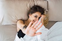 Молода жінка лежить на ліжку з ковдрою і витягнутою рукою, щоб покрити і сміятися — стокове фото