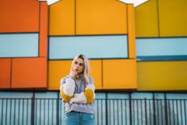 Mujer bonita joven reflexiva de pie contra casas coloridas y mirando hacia otro lado - foto de stock