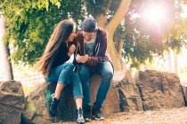 Смеющаяся молодая пара сидит на скале со смартфоном в парке — стоковое фото