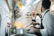 Excited cook making a flambe in restaurant kitchen with colleague watching — Stock Photo