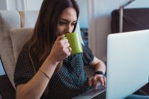Mulher usando laptop em poltrona e segurando xícara de chá — Fotografia de Stock