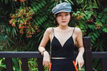 Joven mujer asiática en ropa elegante de pie en la cerca en el parque y mirando a la cámara - foto de stock