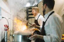 Cocinero emocionado haciendo un flambe en la cocina del restaurante con colega viendo - foto de stock
