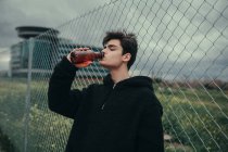 Joven hombre guapo bebiendo de botella de plástico en la valla - foto de stock