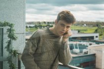 Blick durch das Glas eines ernsten jungen Mannes im Pullover, der sich auf dem Balkon an die Hand lehnt und in die Kamera blickt — Stockfoto