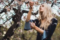 Junge blonde Frau steht am blühenden Baum und berührt Blüte — Stockfoto