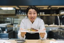 Улыбающийся шеф-повар показывает тарелку с едой в ресторане — стоковое фото