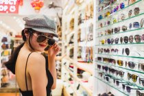 Портрет стильний азіатських брюнетки стоячи в магазині і намагається сонцезахисні окуляри — стокове фото
