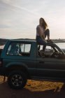 Женщина сидит на крыше автомобиля на берегу и смотрит в сторону — стоковое фото