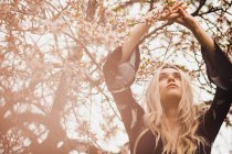 Young blonde woman standing at blooming tree with hands up — Stock Photo