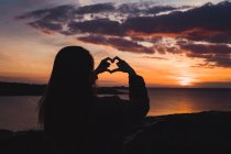 Silhouette of woman standing at ocean at sunset and gesturing heart with fingers — Stock Photo