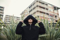 Uomo con cappuccio nero giacca in piedi nel cespuglio verde in città — Foto stock
