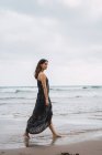 Elegante donna in abito nero passeggiando sulla spiaggia — Foto stock