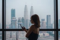 Jolie femme utilisant un smartphone à la fenêtre dans un appartement avec vue sur la ville — Photo de stock