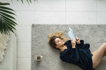 Жінка з кавою і книгою лежить на килимі в сучасній квартирі — стокове фото