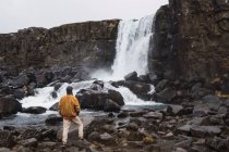 Homem de pé com as mãos em bolsos na frente do fluxo de cachoeira, Islândia — Fotografia de Stock