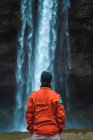 Uomo in cappotto invernale arancione in piedi con le mani in tasca davanti al ruscello della cascata, Islanda — Foto stock