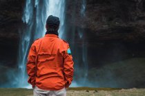 Homme en manteau d'hiver orange debout avec les mains dans les poches devant le ruisseau de cascade, Islande — Photo de stock
