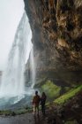Пара, стоящая на холме под скалой с прекрасным водопадом, Исландия — стоковое фото