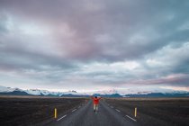 Чоловік у червоній куртці, що йде по дорозі з горами та драматичним небом на фоні — стокове фото