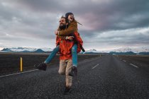 Homme barbu portant femme sur le dos courir et rire sur la route vide près des montagnes — Photo de stock