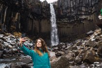Улыбающаяся молодая женщина делает селфи водопад и камни со смартфоном — стоковое фото