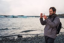 Happy bearded traveler with backpack taking photo with smartphone while standing on pebbled beach, Iceland — Stock Photo