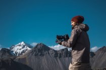Uomo barbuto tenendo fotocamera professionale mentre in piedi alla luce del sole delle montagne — Foto stock