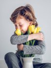 Junge im Grundalter mit geschlossenen Augen sitzt und umarmt ein Bündel gelber Tulpen auf grauem Hintergrund. — Stockfoto