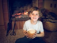 Fröhlicher Junge sitzt mit Tasse am Kamin — Stockfoto