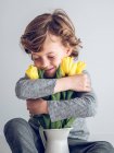 Elementary age boy with eyes closed sitting and embracing bunch of yellow tulips on gray background. — Stock Photo