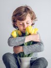 Niño de edad elemental con los ojos cerrados sentado y abrazando racimo de tulipanes amarillos sobre fondo gris . - foto de stock