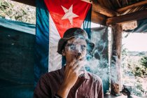 LA HABANA, CUBA - 1 MAGGIO 2018: Uomo in cappello fuma sigaro spesso in piedi contro la bandiera di Cuba in una piccola capanna alla luce del sole — Foto stock