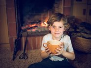 Веселий хлопчик сидить біля каміна з чашкою — стокове фото