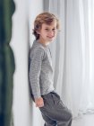 Alegre elementar idade menino inclinado no parede e olhando para câmera dentro de casa . — Fotografia de Stock