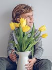 Хлопчик початкового віку з жовтими тюльпанами в глечику дивиться на сірий фон . — стокове фото