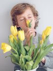 Elementary age boy smelling yellow tulips in jug against gray background. — Stock Photo