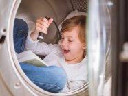 Menino idade elementar com lanterna deitado dentro da máquina de lavar roupa com livro . — Fotografia de Stock