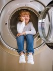 Menino da idade elementar sentado dentro da máquina de lavar roupa com a mão no queixo . — Fotografia de Stock
