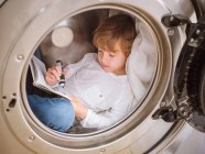 Menino da idade elementar com lanterna deitado dentro da máquina de lavar roupa e livro de leitura . — Fotografia de Stock