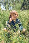 Menino da idade elementar sentado no campo de flores silvestres e segurando planta . — Fotografia de Stock