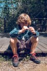 Menino da idade elementar com cabelo louro encaracolado sentado na ponte de madeira e olhando na câmera no campo . — Fotografia de Stock